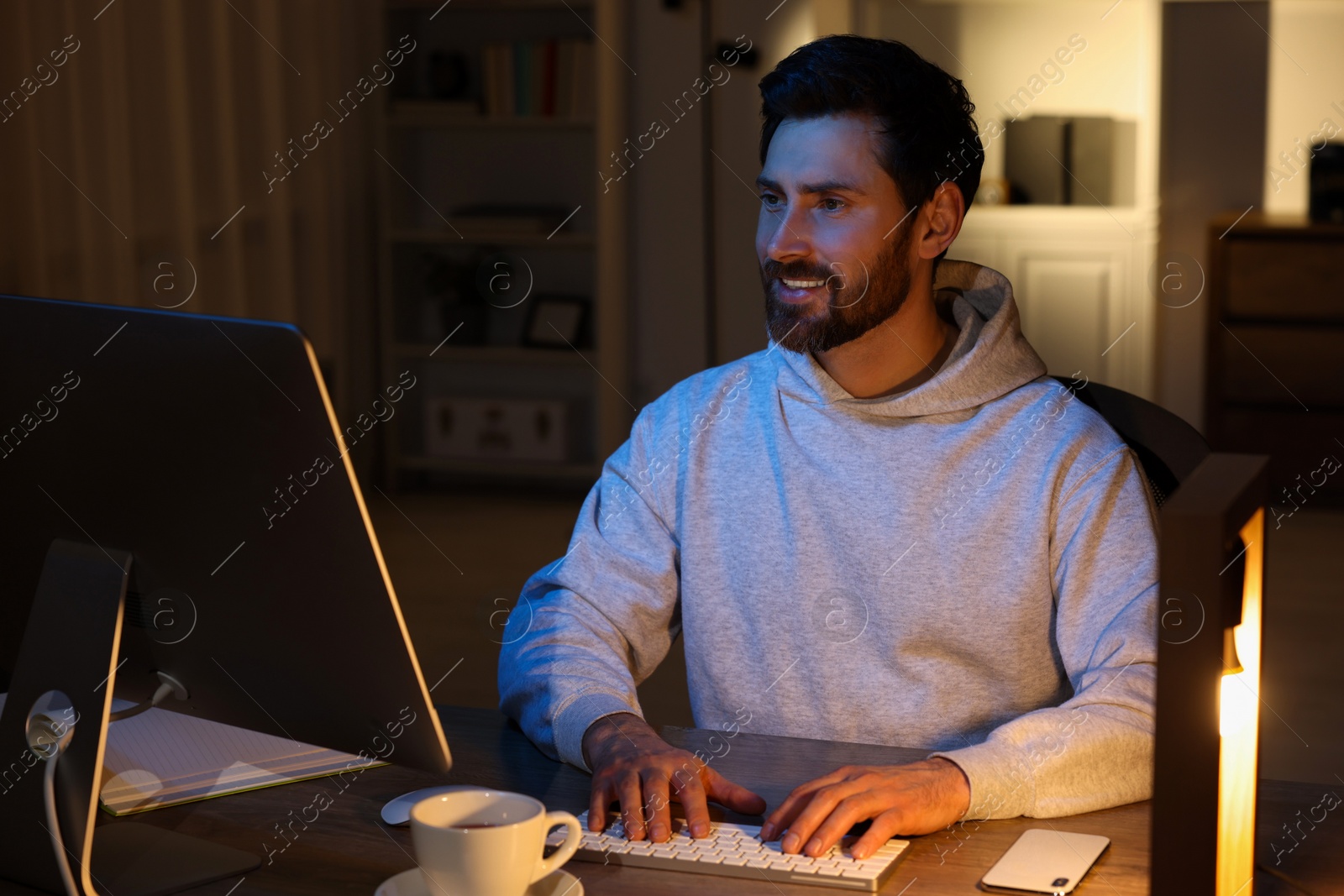 Photo of Home workplace. Happy man working with computer at wooden desk in room at night