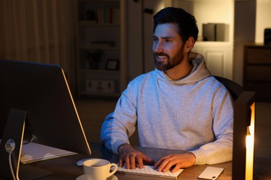 Photo of Home workplace. Happy man working with computer at wooden desk in room at night