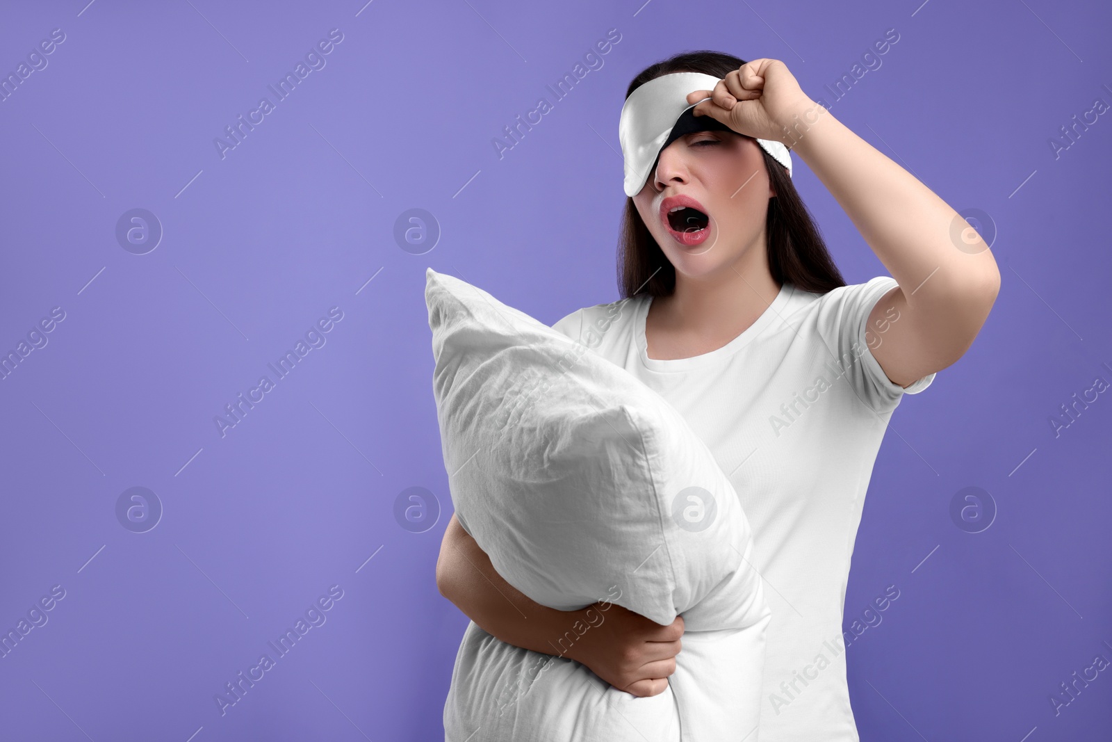 Photo of Tired young woman with sleep mask and pillow yawning on purple background, space for text. Insomnia problem