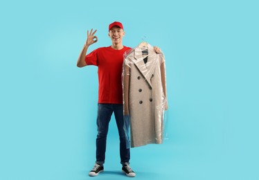 Dry-cleaning delivery. Happy courier holding coat in plastic bag and showing OK gesture on light blue background