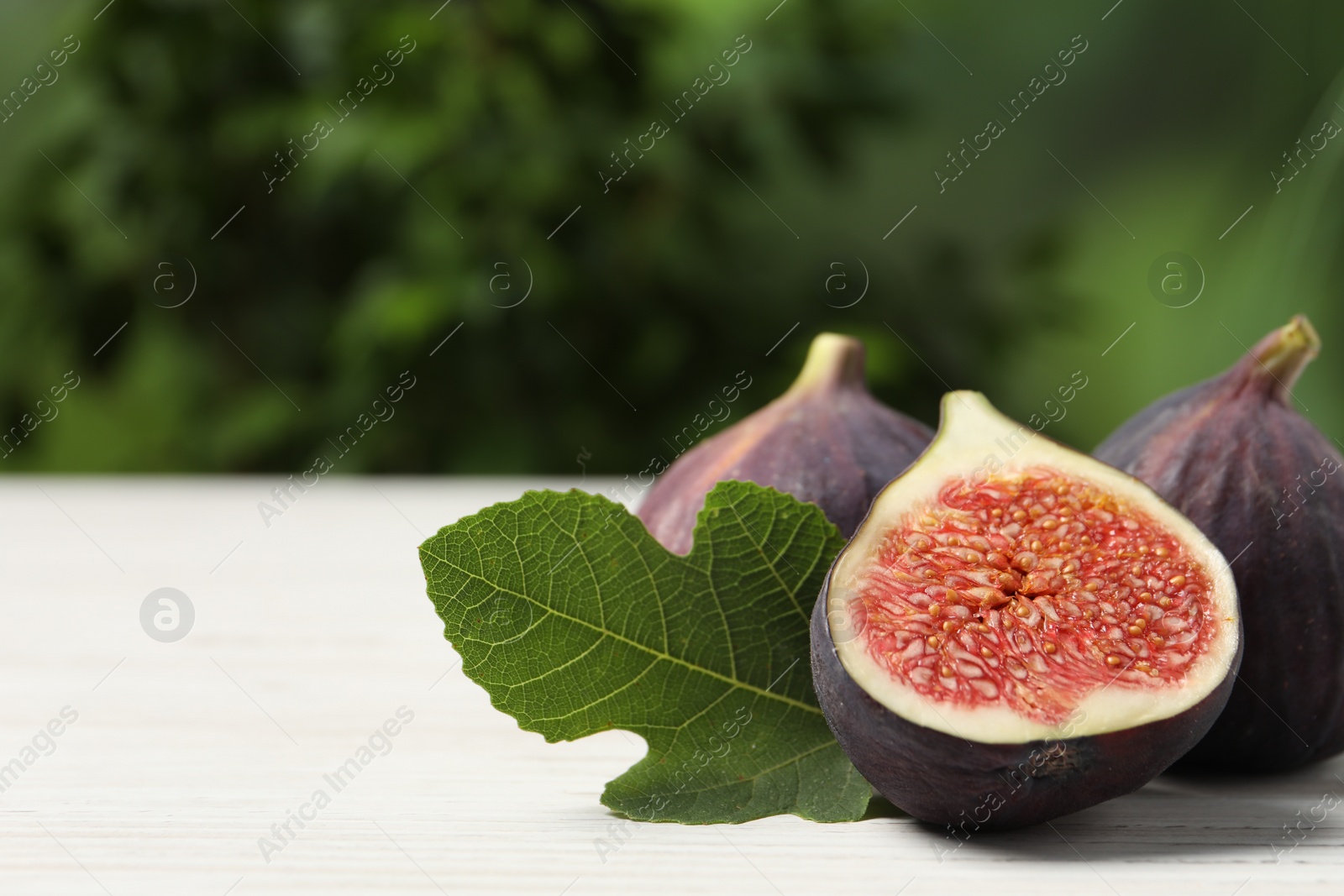 Photo of Whole and cut ripe figs with leaf on white wooden table against blurred green background, closeup. Space for text