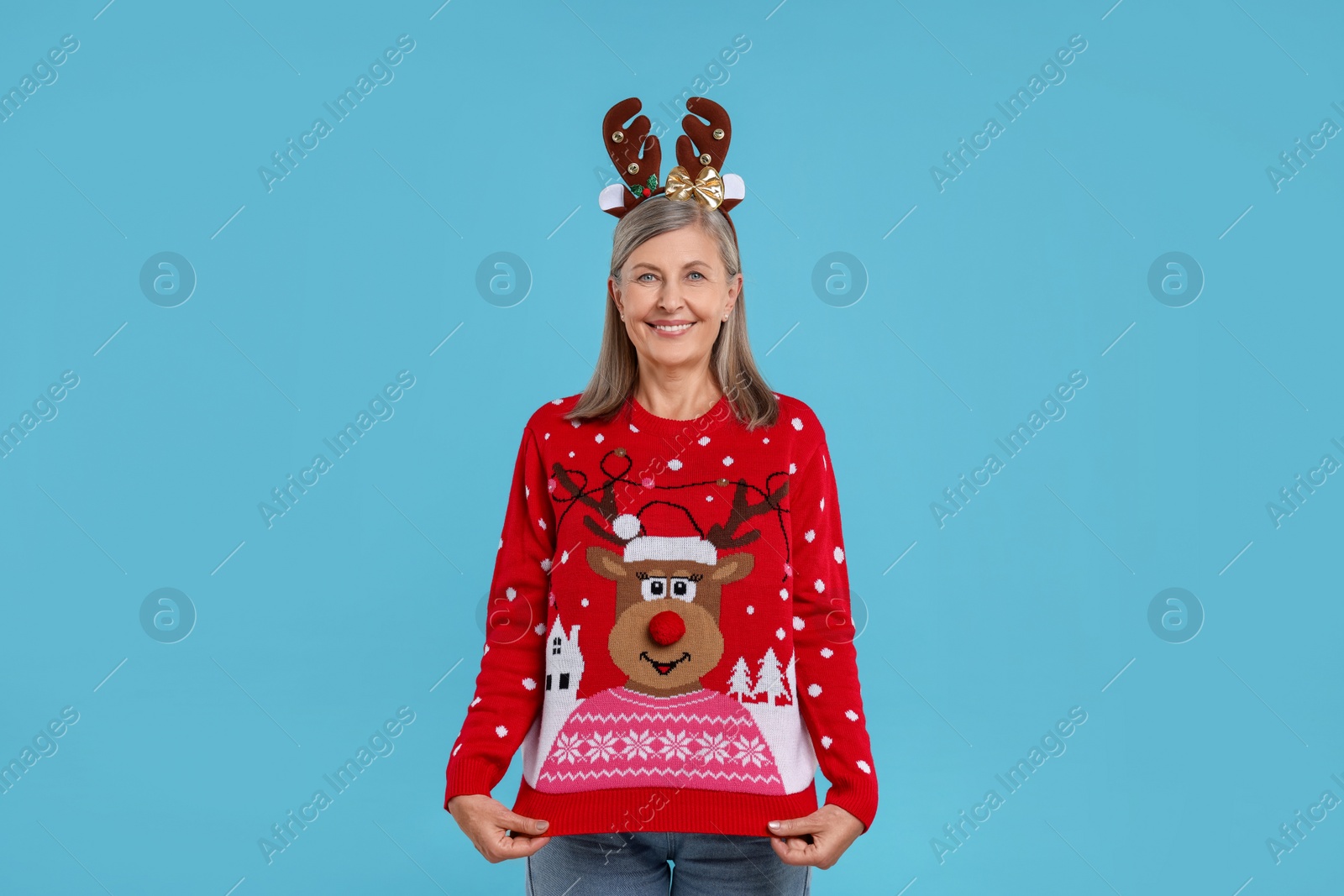 Photo of Happy senior woman in reindeer headband showing Christmas sweater on light blue background