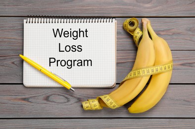 Photo of Notebook with phrase Weight Loss Program, measuring tape and bananas on grey wooden table, flat lay
