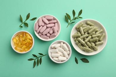 Photo of Different vitamin capsules in bowls and leaves on turquoise background, flat lay