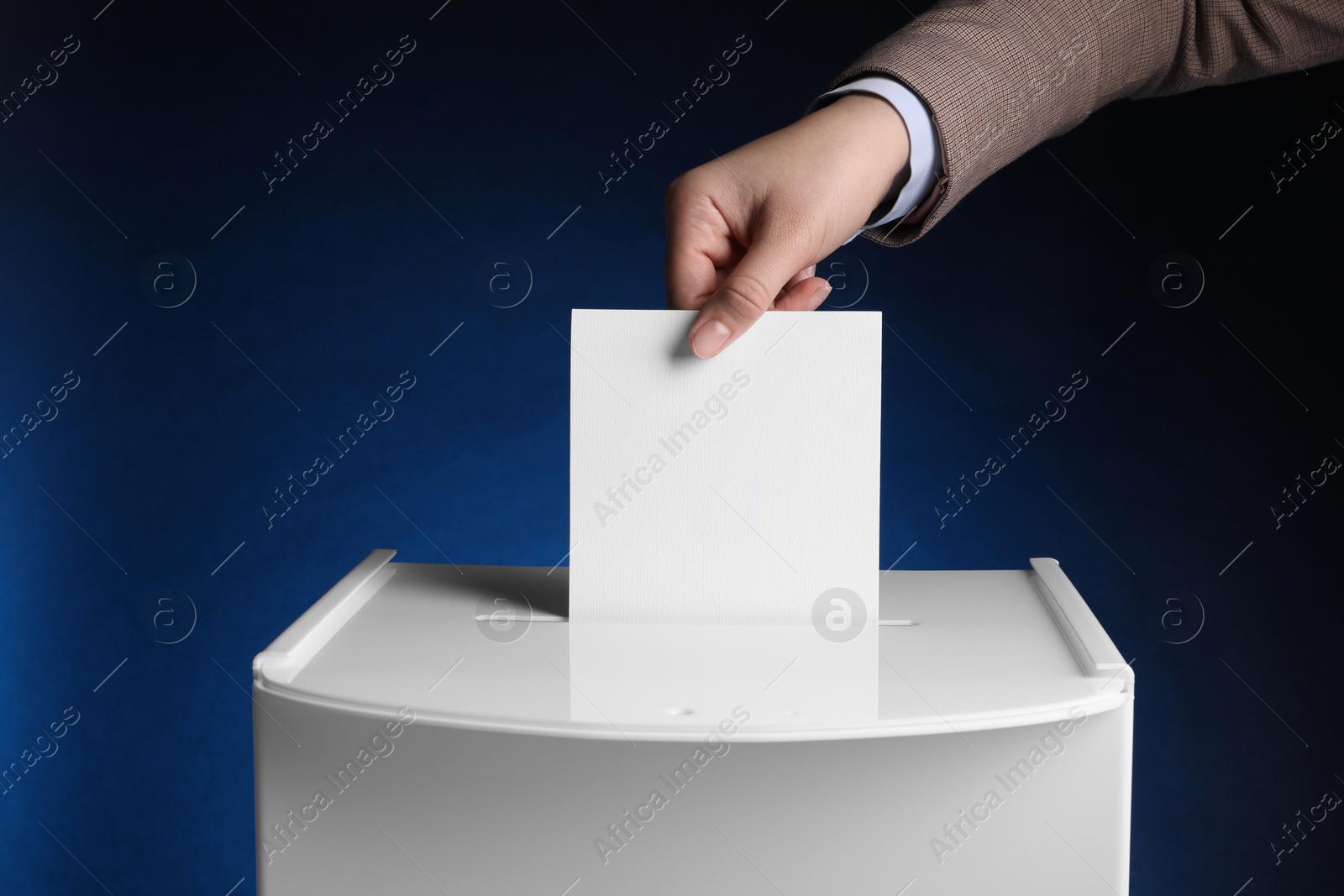 Photo of Woman putting her vote into ballot box on dark blue background, closeup