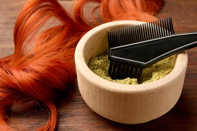 Bowl of henna powder, brush and red strand on wooden table, closeup. Natural hair coloring