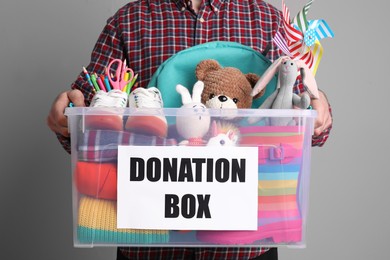 Photo of Man holding donation box full of different toys, clothes and stationery on grey background, closeup
