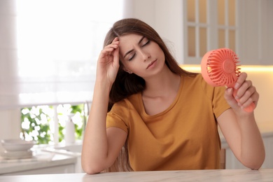 Photo of Woman with portable fan at table in kitchen. Summer heat