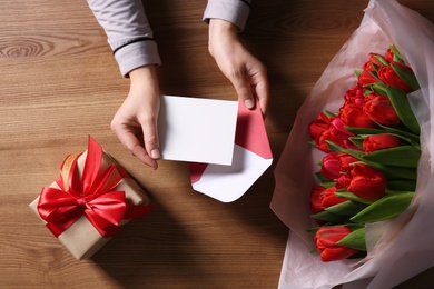 Photo of Woman holding envelope with blank greeting card, bouquet of tulips and gift box on wooden table, top view