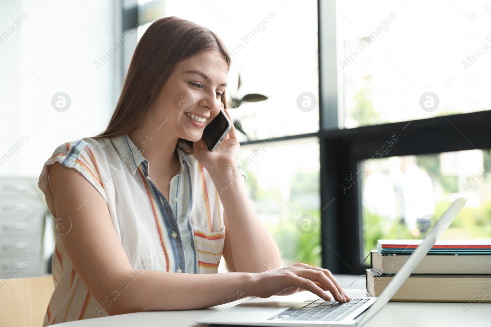 Photo of Young woman talking on phone and working with laptop at table in library