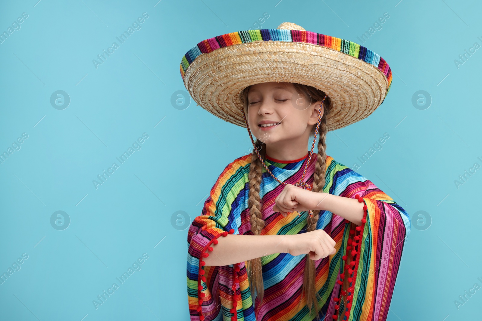 Photo of Cute girl in Mexican sombrero hat and poncho dancing on light blue background. Space for text