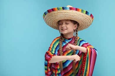 Photo of Cute girl in Mexican sombrero hat and poncho dancing on light blue background. Space for text