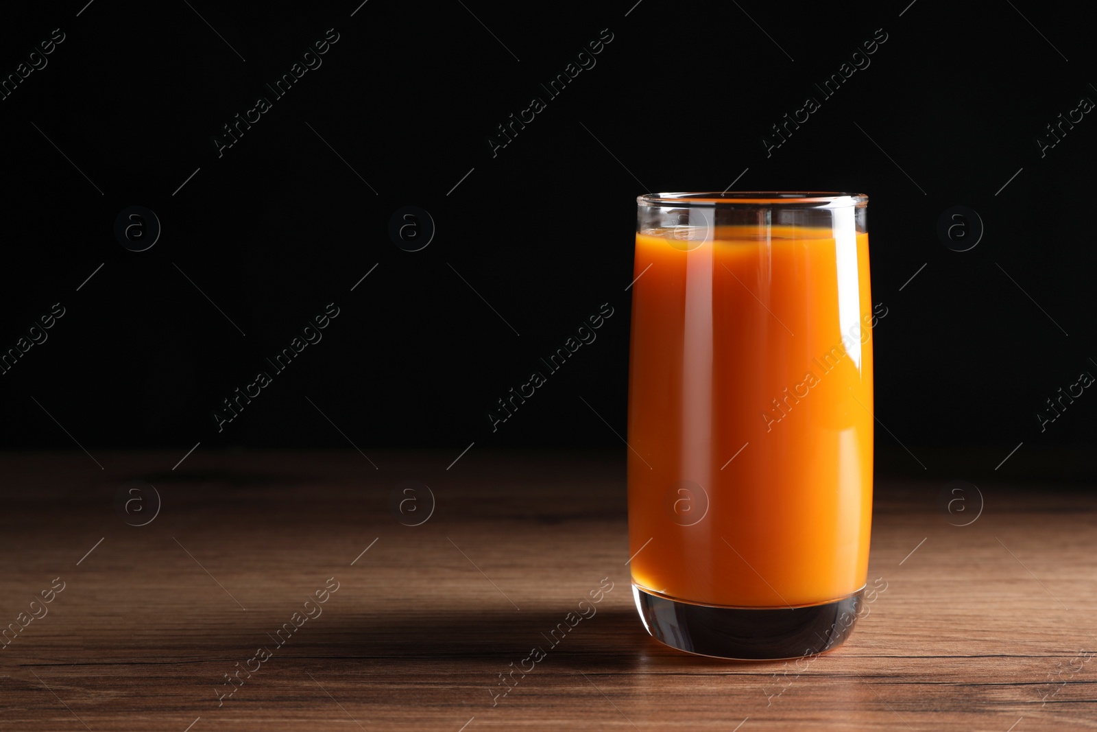 Photo of Glass of tasty carrot juice on wooden table against black background, space for text