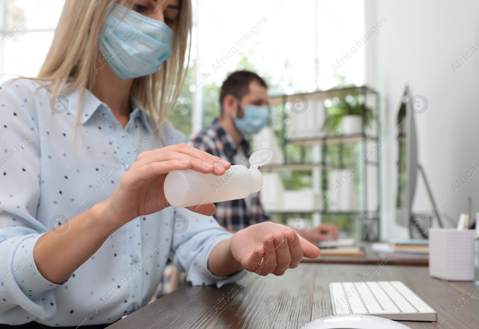 Photo of Office employee in mask applying hand sanitizer at workplace