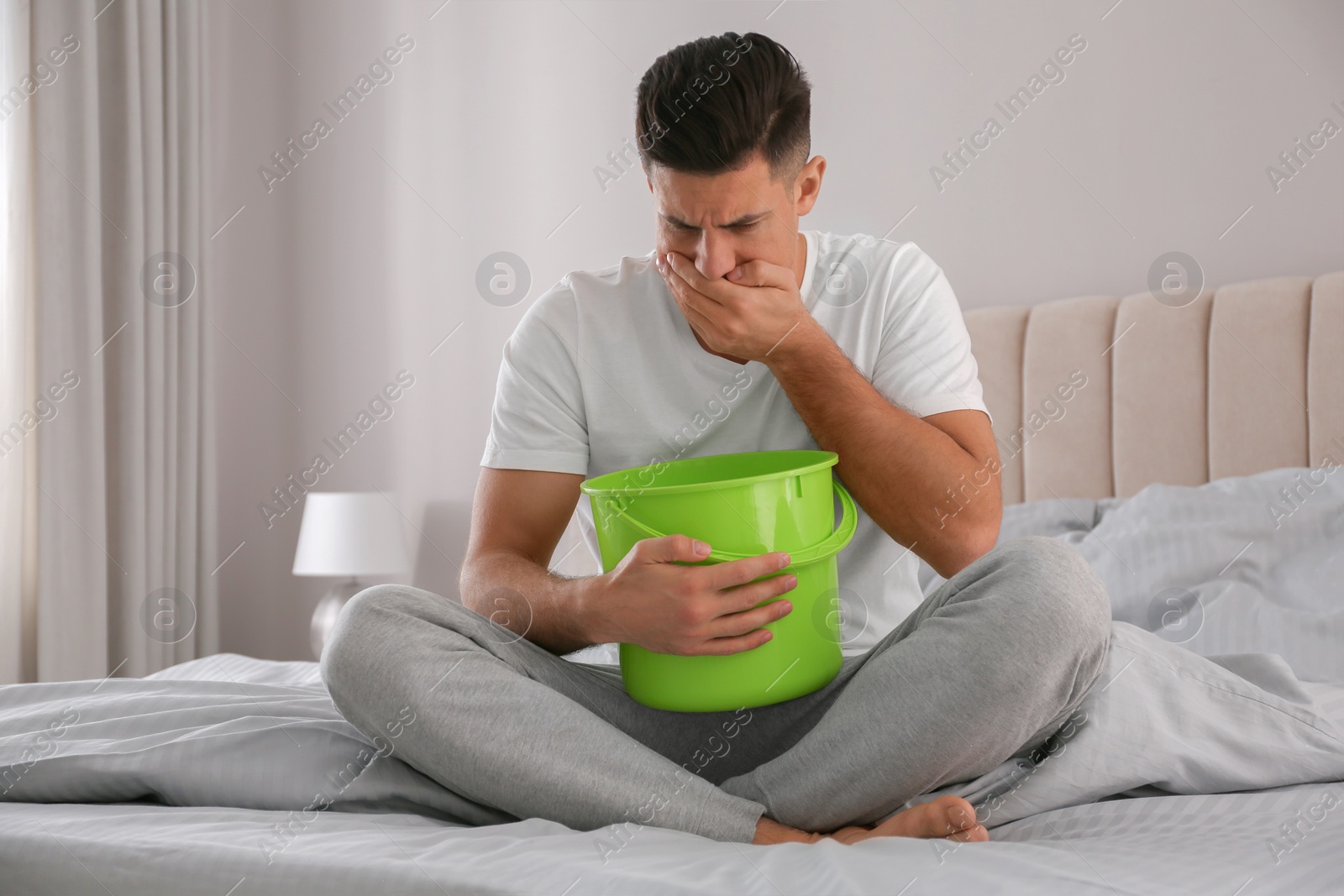 Photo of Man with bucket suffering from nausea on bed at home. Food poisoning