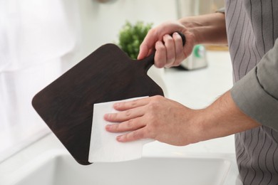 Man wiping dark wooden cutting board with paper napkin at sink in kitchen, closeup