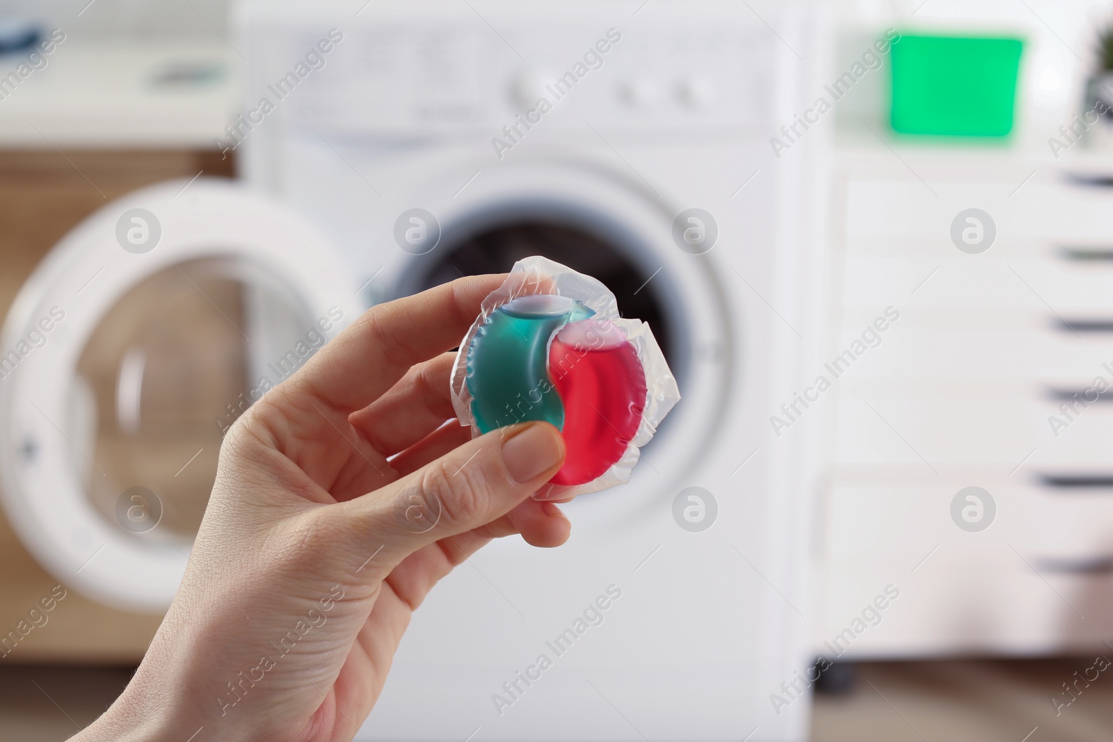 Photo of Woman holding laundry detergent capsule near washing machine indoors, closeup