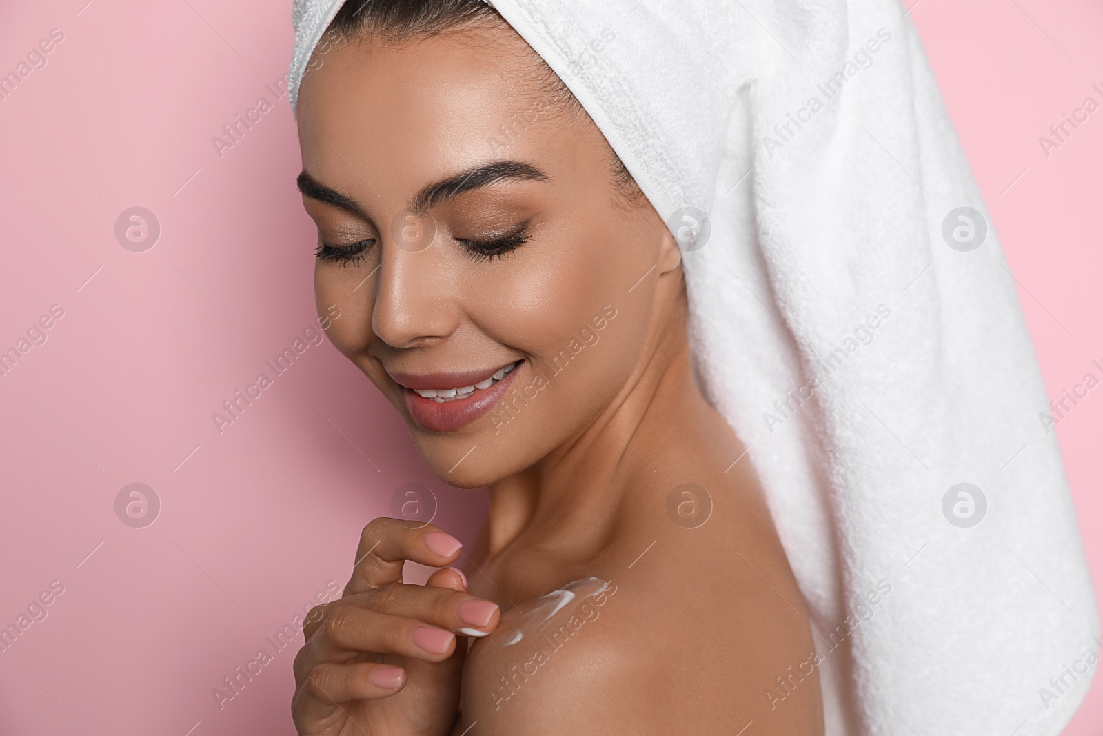 Photo of Beautiful young woman with towel applying cream on shoulder against pink background