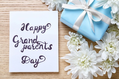 Photo of Beautiful white flowers, gift box and card with phrase Happy Grandparents Day on wooden background, flat lay