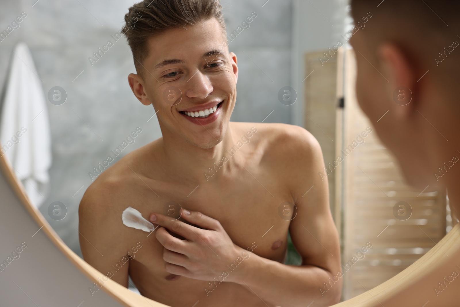 Photo of Handsome man applying moisturizing cream onto his shoulder in bathroom