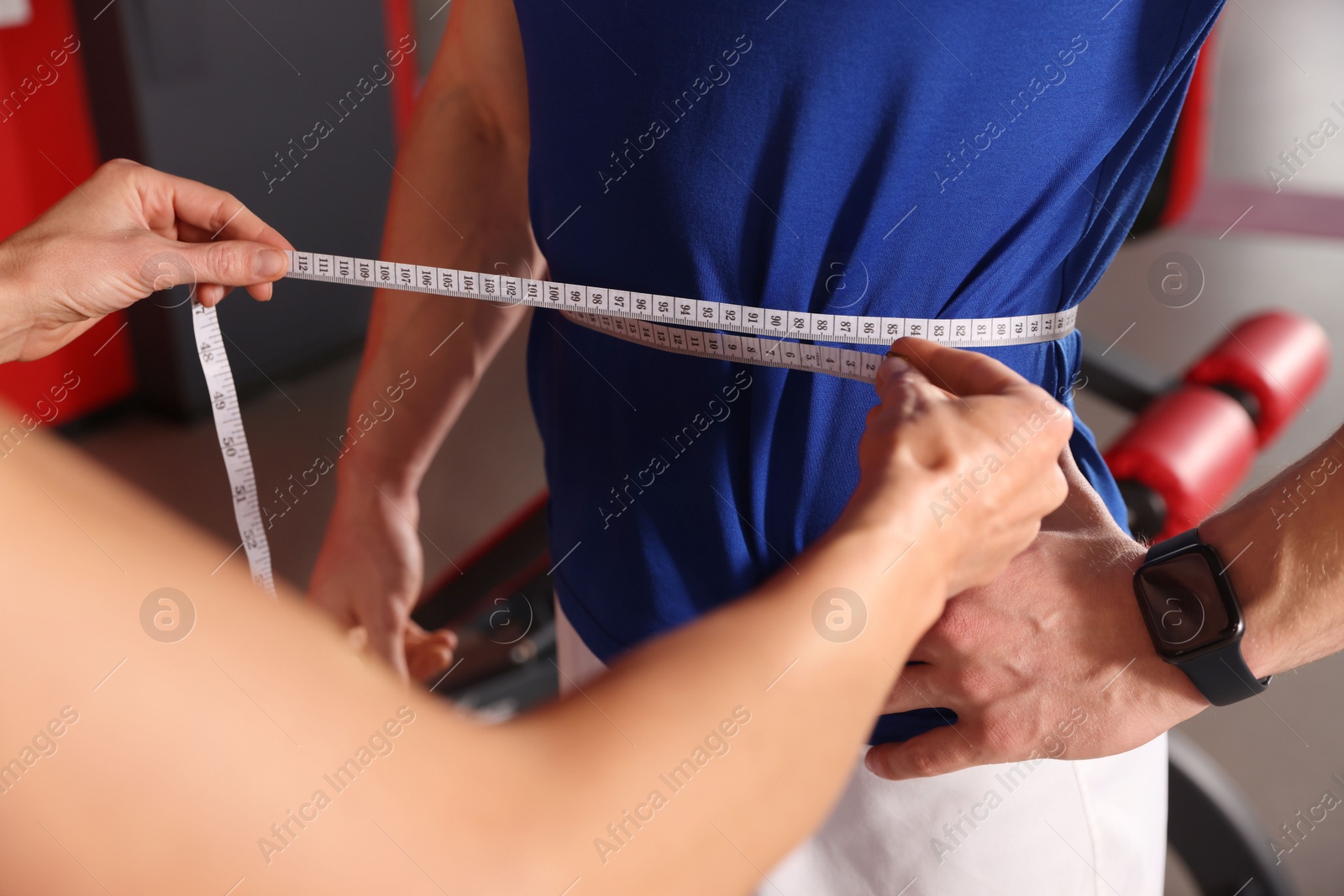 Photo of Trainer measuring man`s waist with tape in gym, closeup