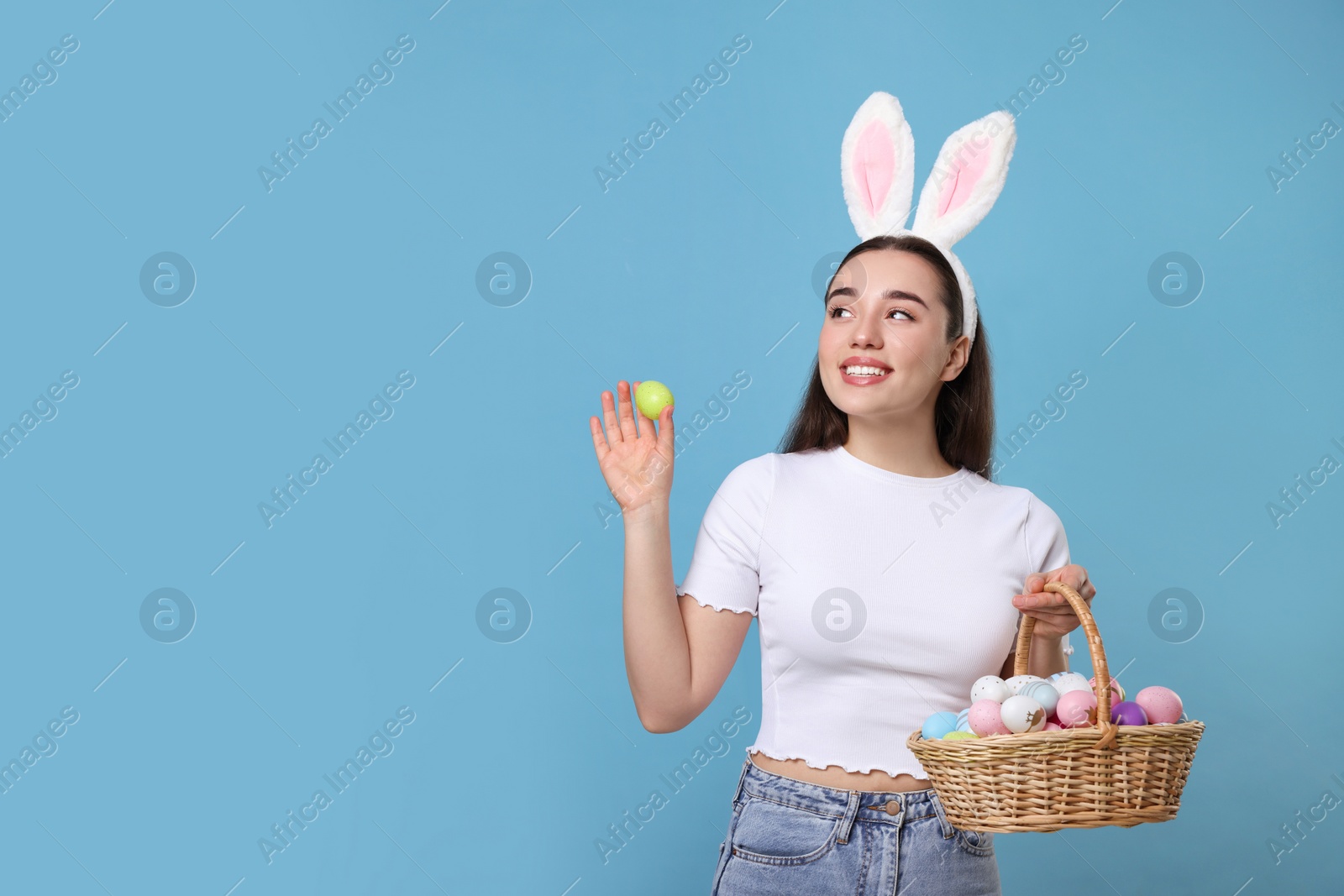 Photo of Happy woman in bunny ears headband holding wicker basket of painted Easter eggs on turquoise background. Space for text