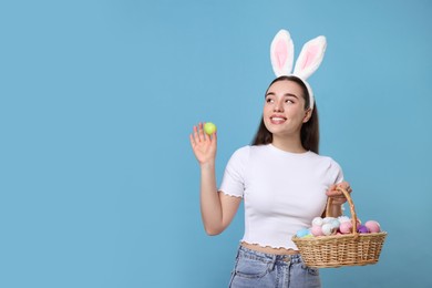 Photo of Happy woman in bunny ears headband holding wicker basket of painted Easter eggs on turquoise background. Space for text
