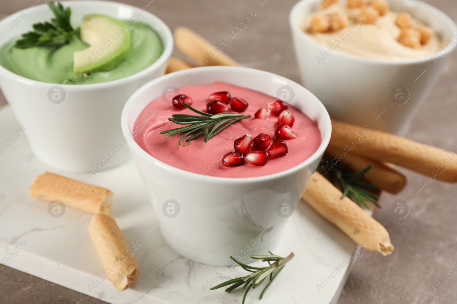 Photo of Bowl of delicious pink hummus with rosemary and pomegranate seeds on brown marble table, closeup