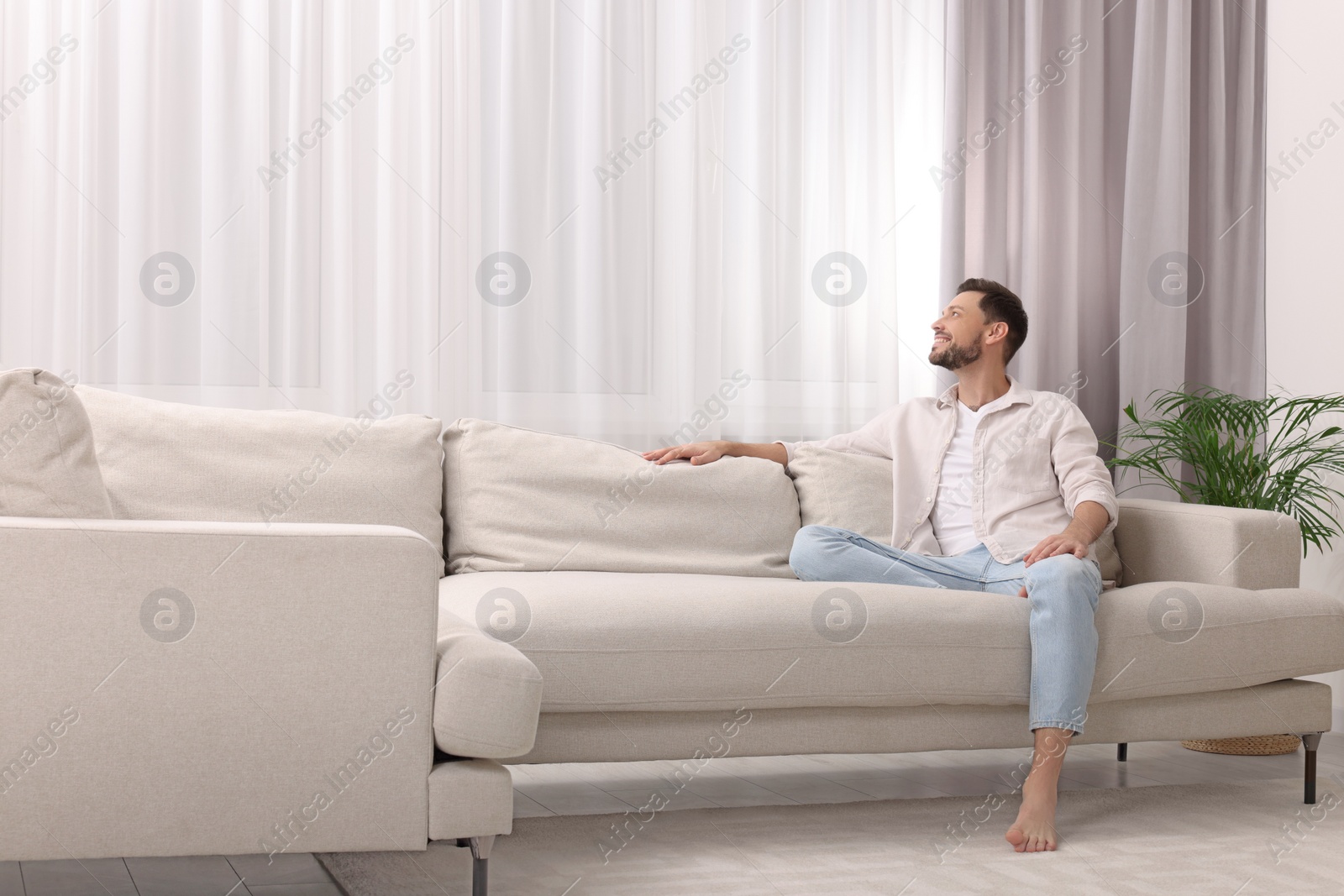 Photo of Happy man on resting sofa near window with beautiful curtains in living room. Space for text