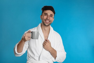 Photo of Young man in bathrobe with cup of coffee on light blue background