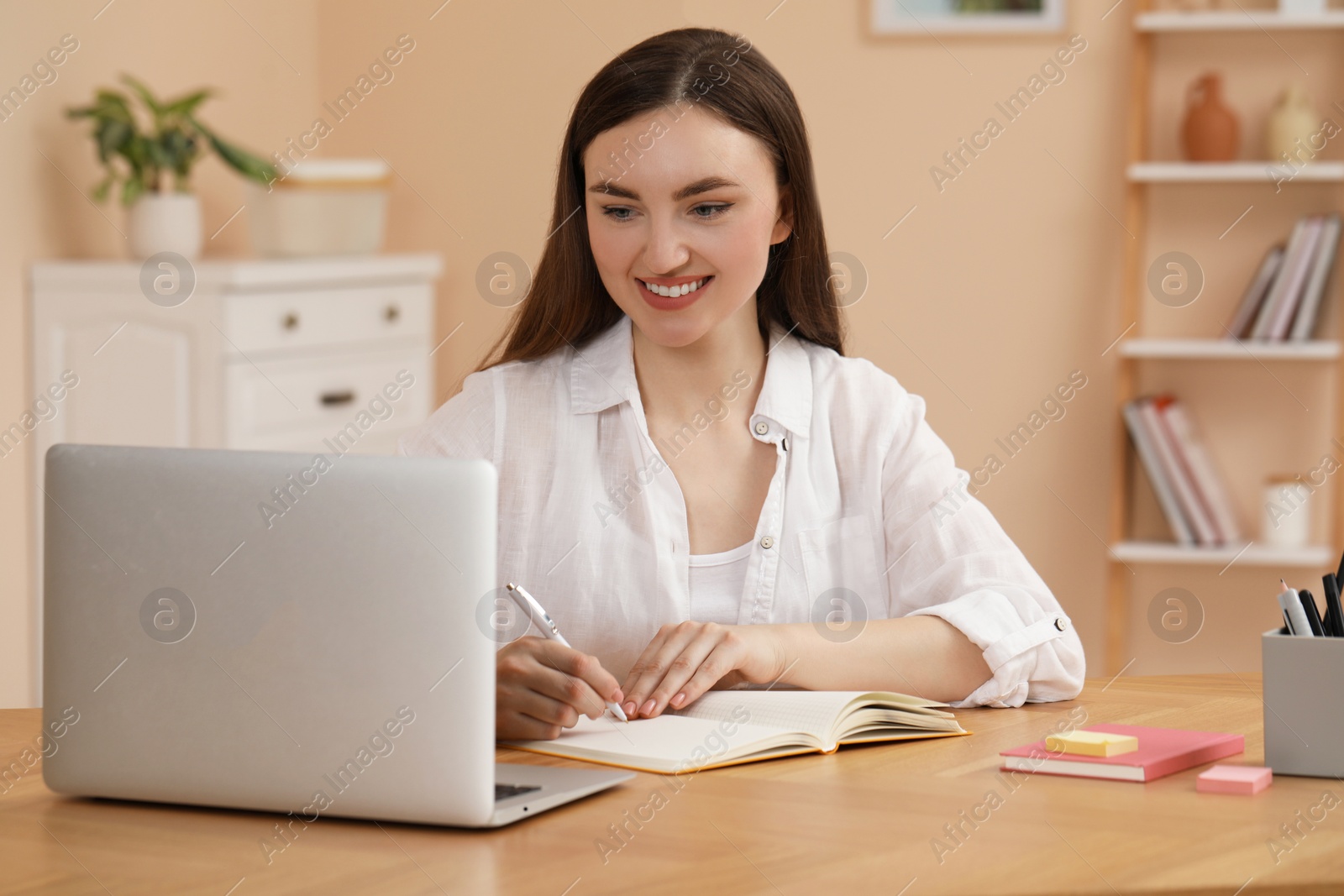 Photo of Happy young woman writing in notebook while working on laptop at wooden table indoors