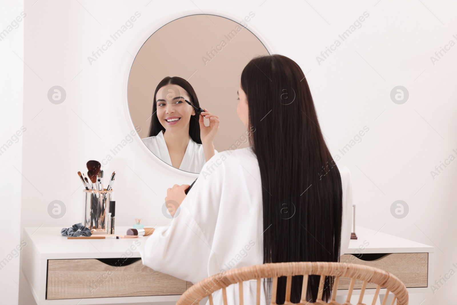 Photo of Beautiful young woman applying mascara at dressing table indoors
