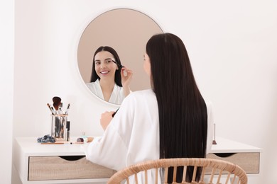Beautiful young woman applying mascara at dressing table indoors
