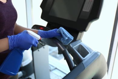 Photo of Woman cleaning treadmill with disinfectant spray and cloth in gym, closeup