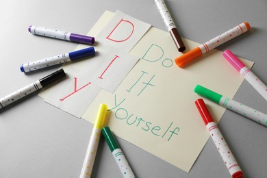 Photo of Sheet with written phrase Do It Yourself and colorful markers on grey background. DIY concept