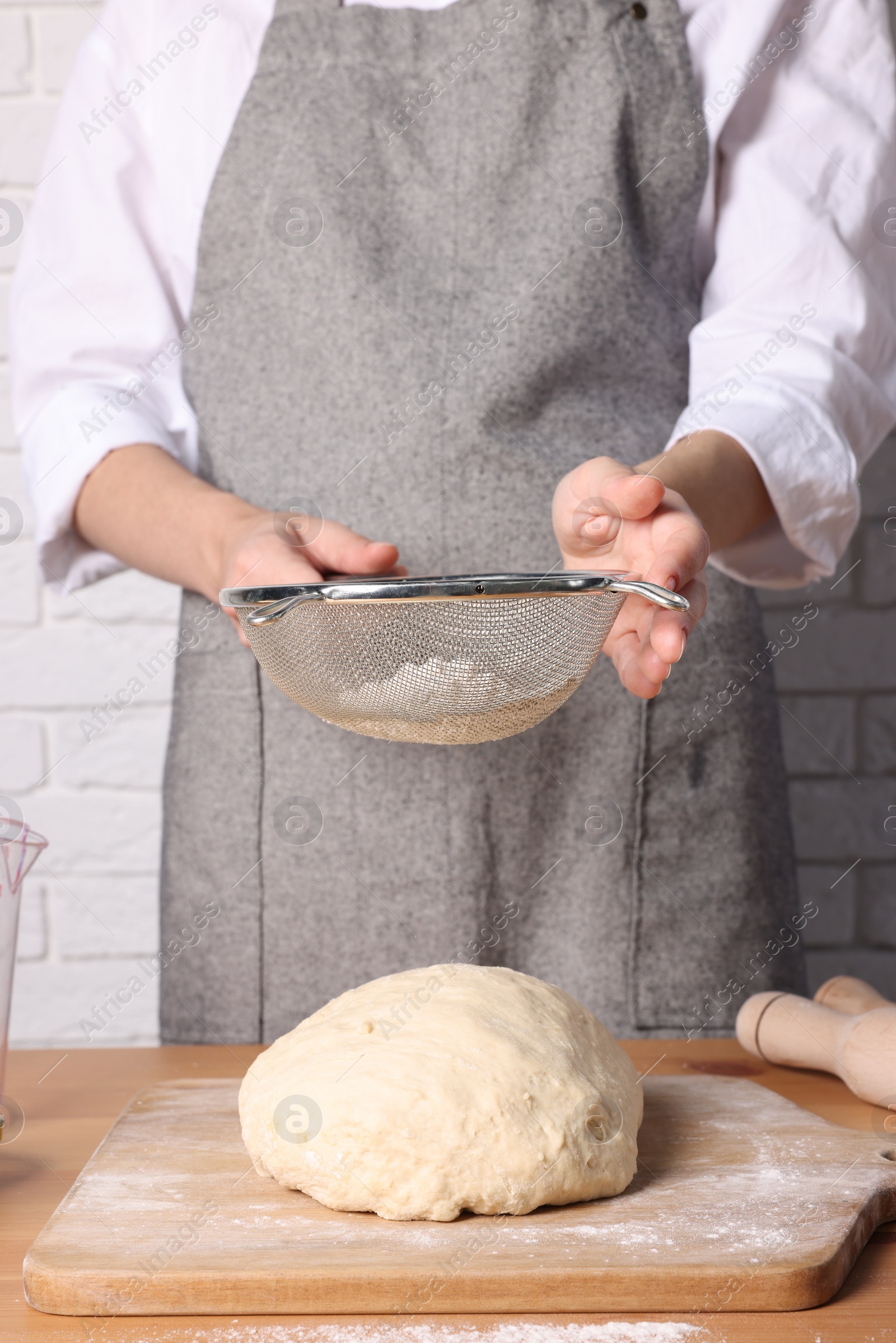 Photo of Woman sprinkling flour over dough at table near white brick wall, closeup