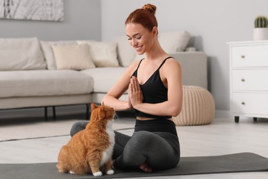 Beautiful woman with cute red cat practicing yoga on mat at home