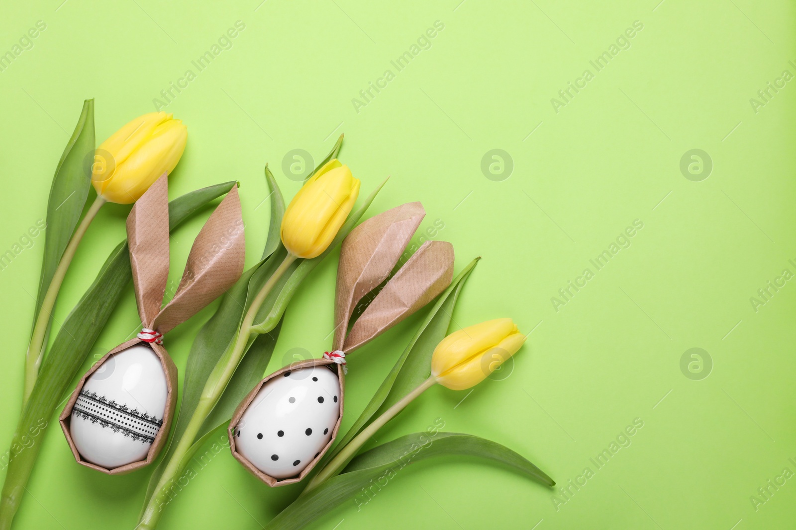 Photo of Easter bunnies made of craft paper and eggs among beautiful tulips on light green background, flat lay. Space for text