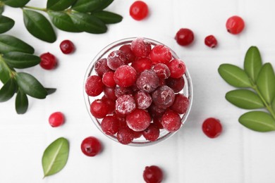 Frozen red cranberries in bowl and green leaves on white table, flat lay