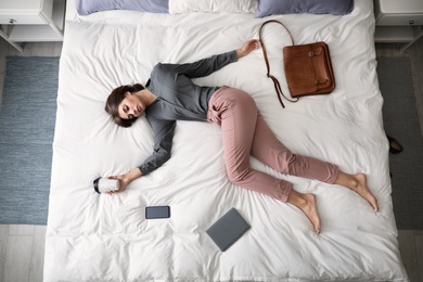 Photo of Exhausted woman with cup of coffee sleeping fully dressed on bed at home, above view