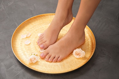 Photo of Woman soaking her feet in plate with water and roses on grey background, closeup. Spa treatment