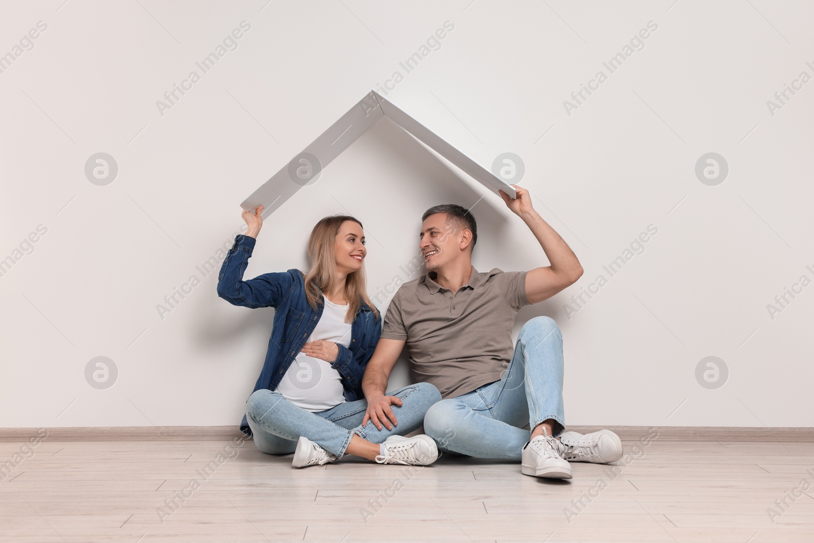 Photo of Young family housing concept. Pregnant woman with her husband sitting under cardboard roof on floor indoors