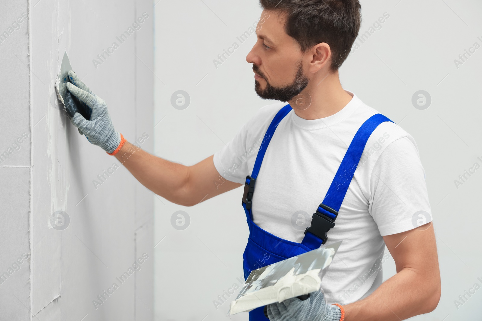 Photo of Professional worker in uniform plastering wall with putty knife indoors