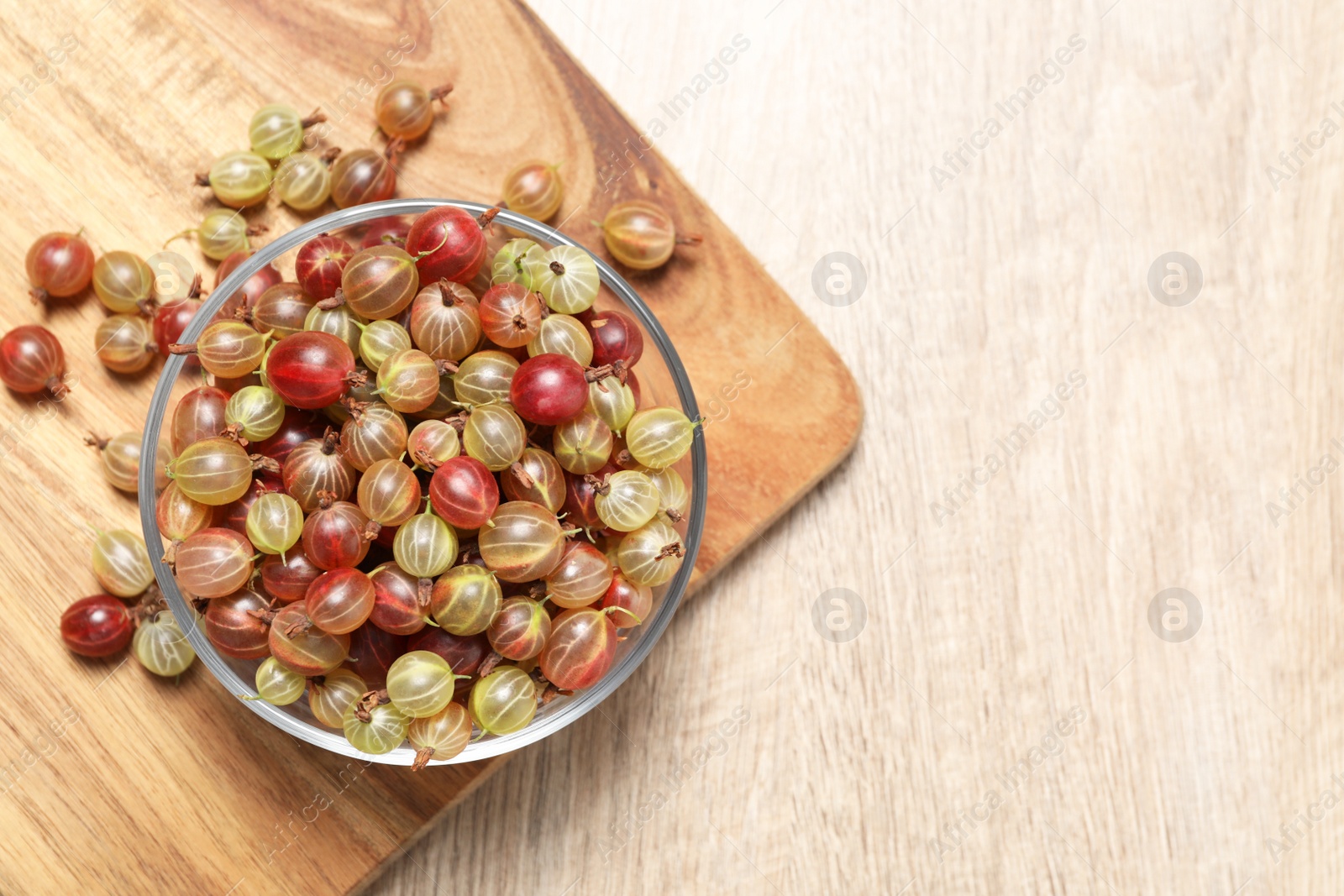 Photo of Glass bowl with fresh ripe gooseberries on wooden table, flat lay. Space for text