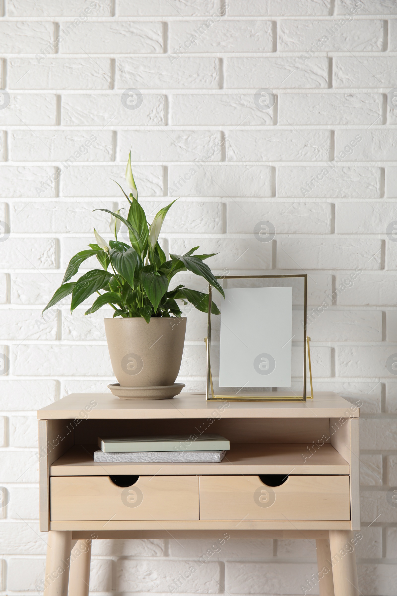 Photo of Spathiphyllum plant in pot and photo frame on table near brick wall, space for text. Home decor