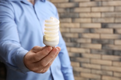Photo of Man holding energy saving bulb for lamp against brick wall, closeup. Space for text