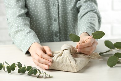 Photo of Furoshiki technique. Woman decorating gift wrapped in fabric with eucalyptus branch at wooden table, closeup