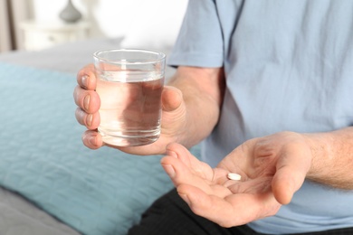 Photo of Senior man holding pill and glass of water indoors, closeup