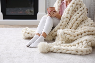 Photo of Woman covered with knitted plaid holding cup of hot drink on floor at home, closeup