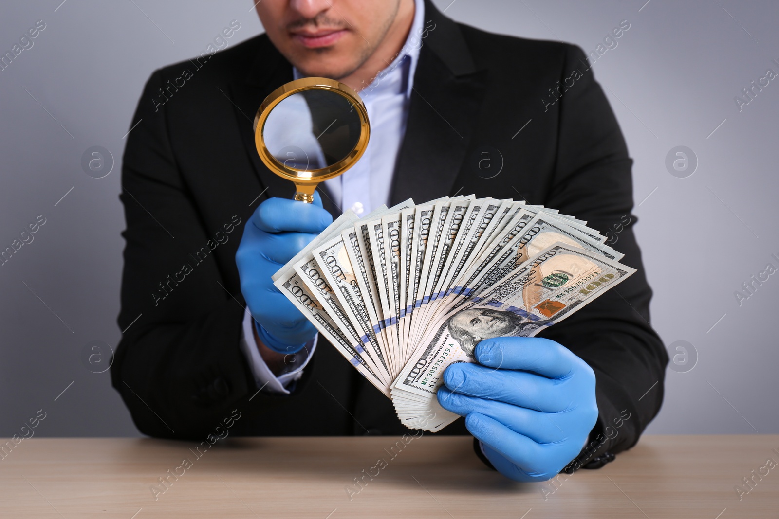 Photo of Expert authenticating 100 dollar banknotes with magnifying glass at table on light grey background, closeup. Fake money concept